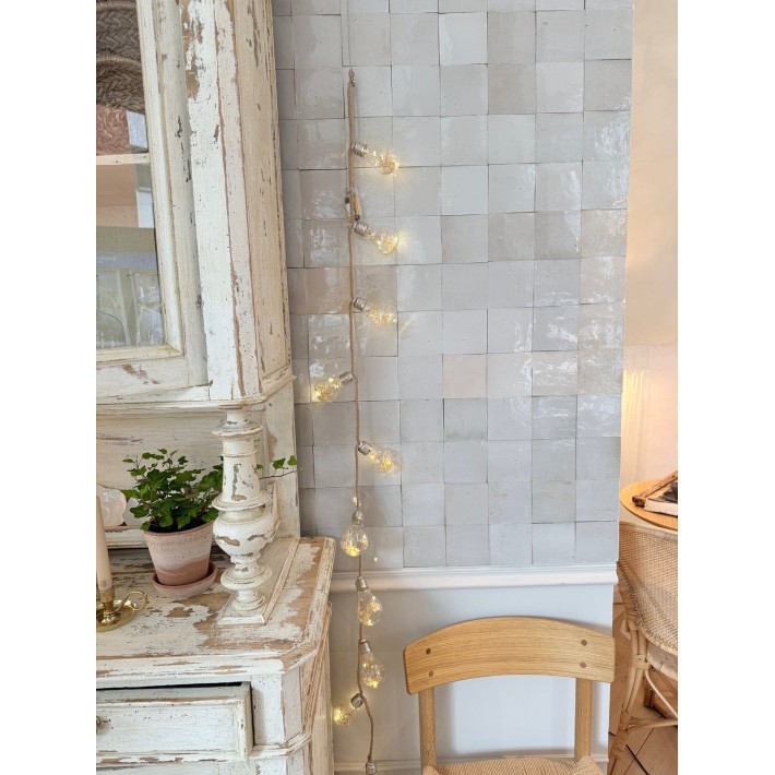 Guirnalda de bombillas decorativas sobre pared de azulejo artesanal, junto a mueble decapado vintage y silla de madera natural