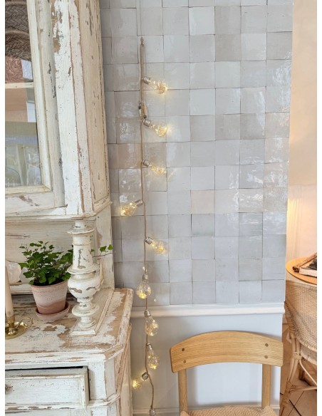 Guirnalda de bombillas decorativas sobre pared de azulejo artesanal, junto a mueble decapado vintage y silla de madera natural Guirnalda de bombillas decorativas sobre pared de azulejo artesanal, junto a mueble decapado vintage y silla de madera natural